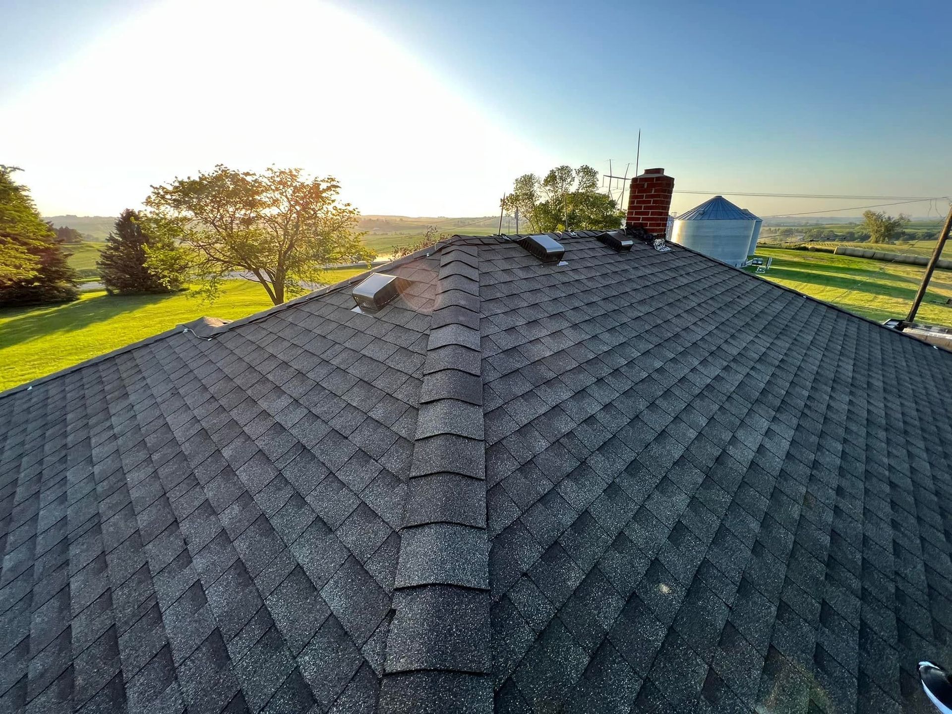 Shingle roof at sunset with chimney, trees, and open fields in the background