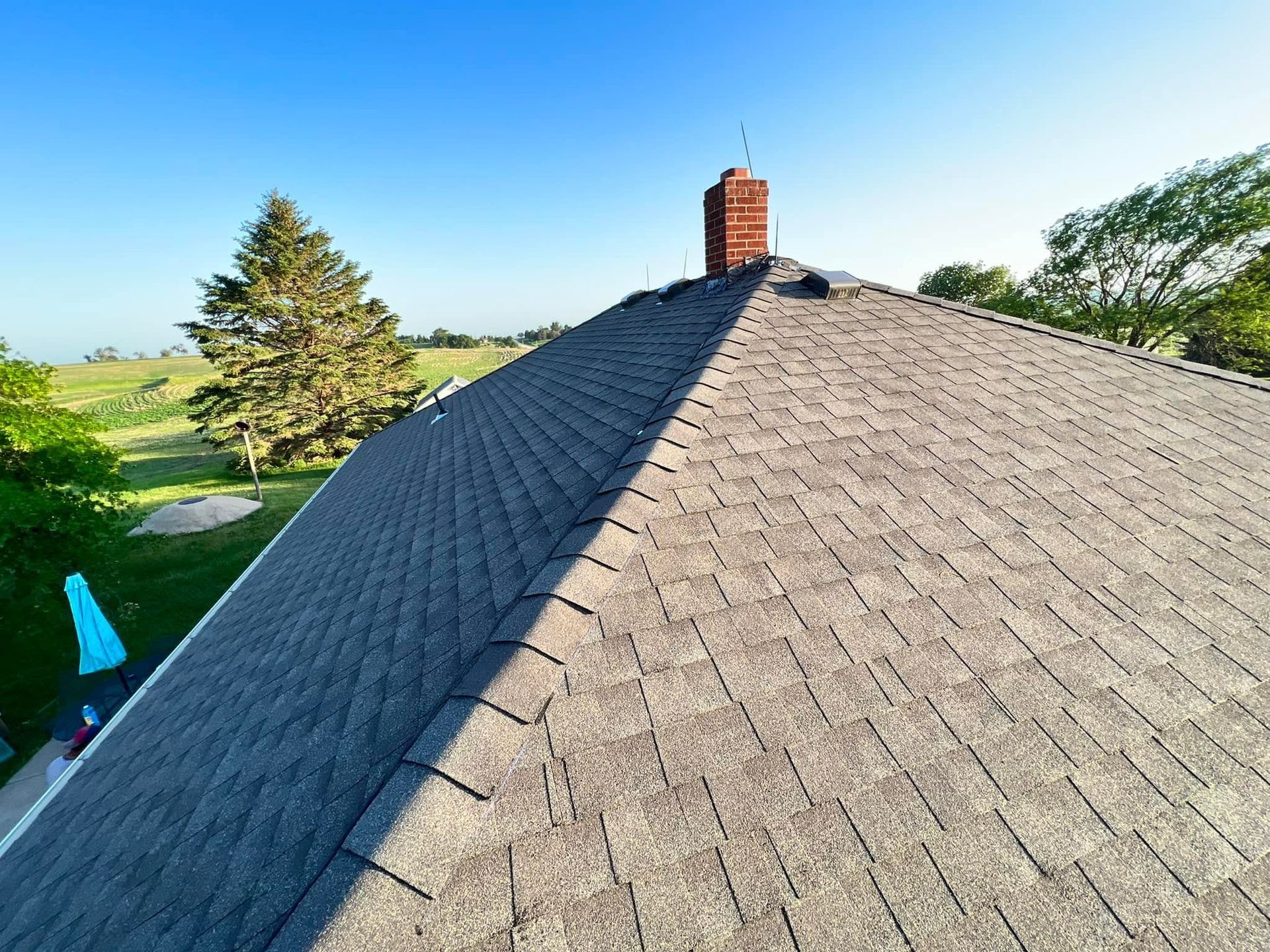 Rooftop with gray shingles and brick chimney, viewed from above with trees and blue sky in the background