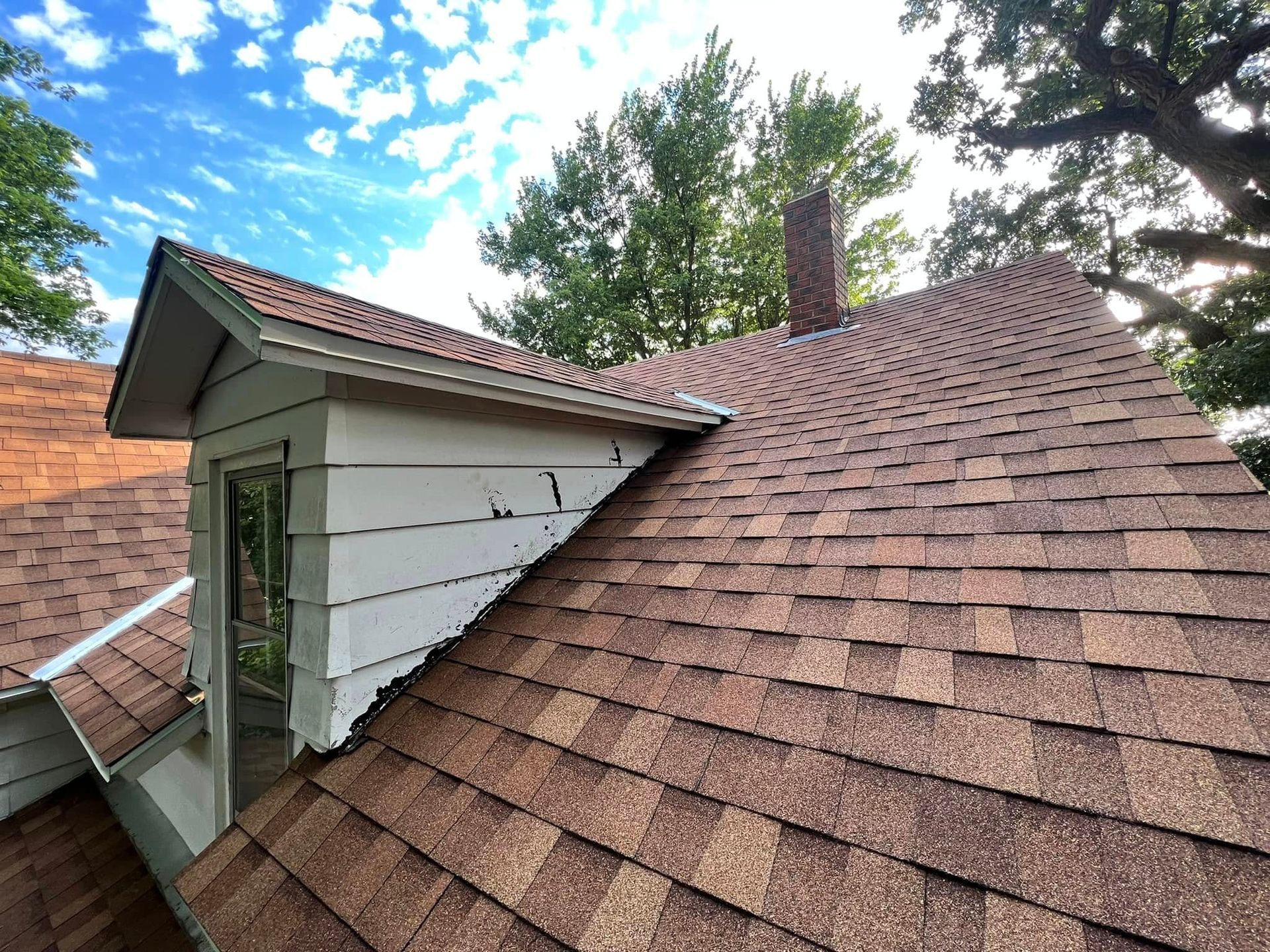 Close-up of a house roof with brown shingles, dormer windows, and blue sky with clouds above