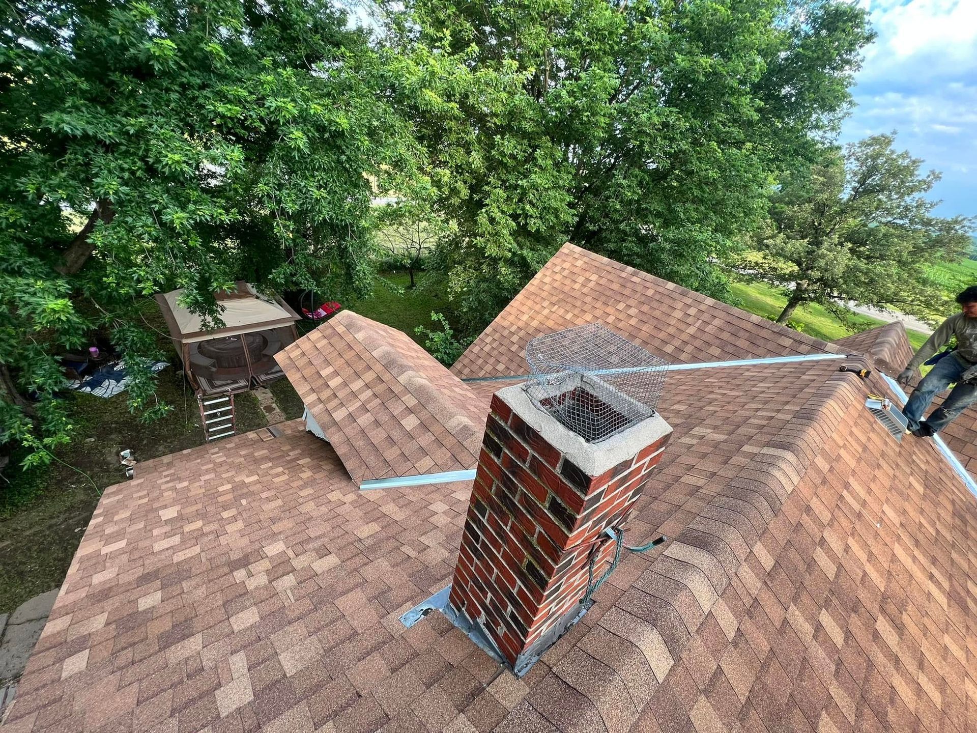 Rooftop with brown shingles, brick chimney, and large green trees surrounding a house