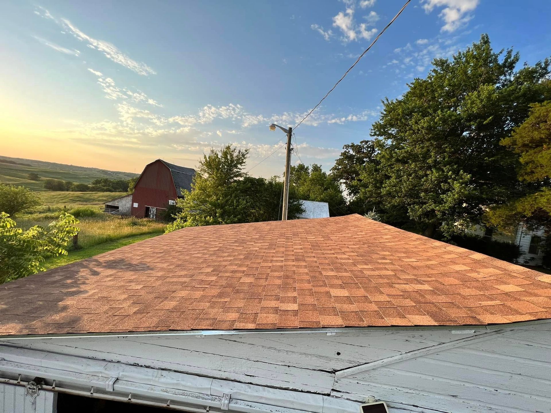 Rural rooftop at sunset with a red barn, trees, and a wide sky over fields