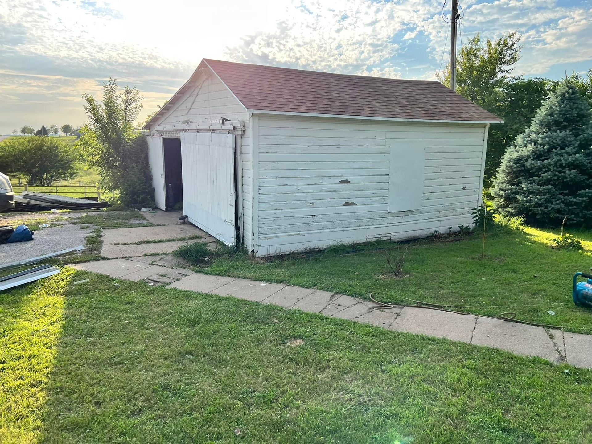 Small white shed with a red roof in a grassy yard, with a concrete walkway and trees behind.