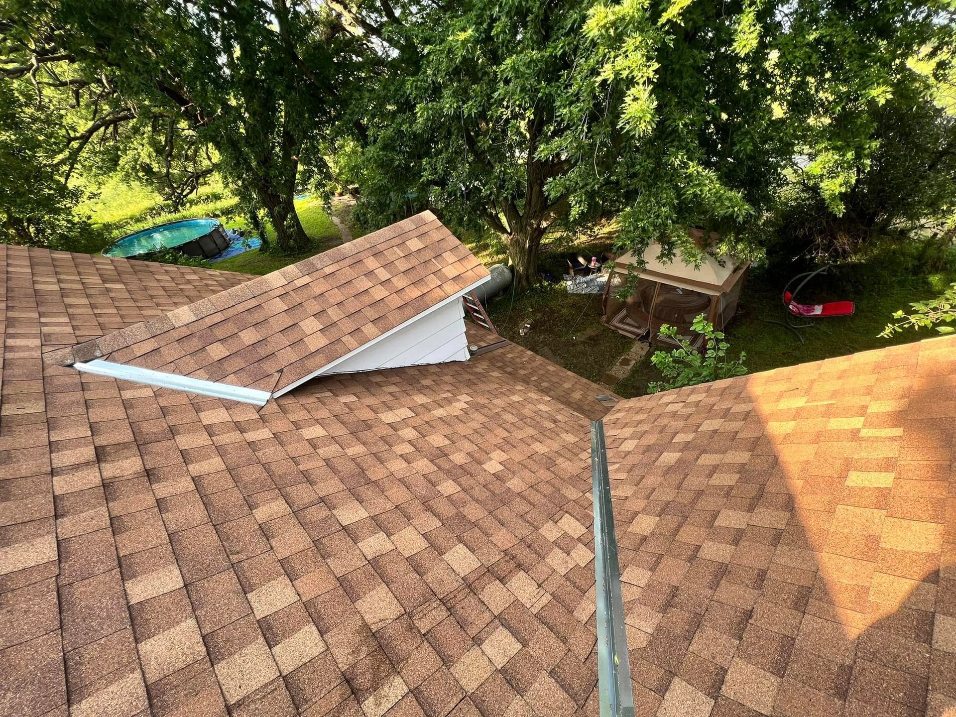 Roof shingles and trees viewed from above, with a damaged vent or panel on the left roof slope.