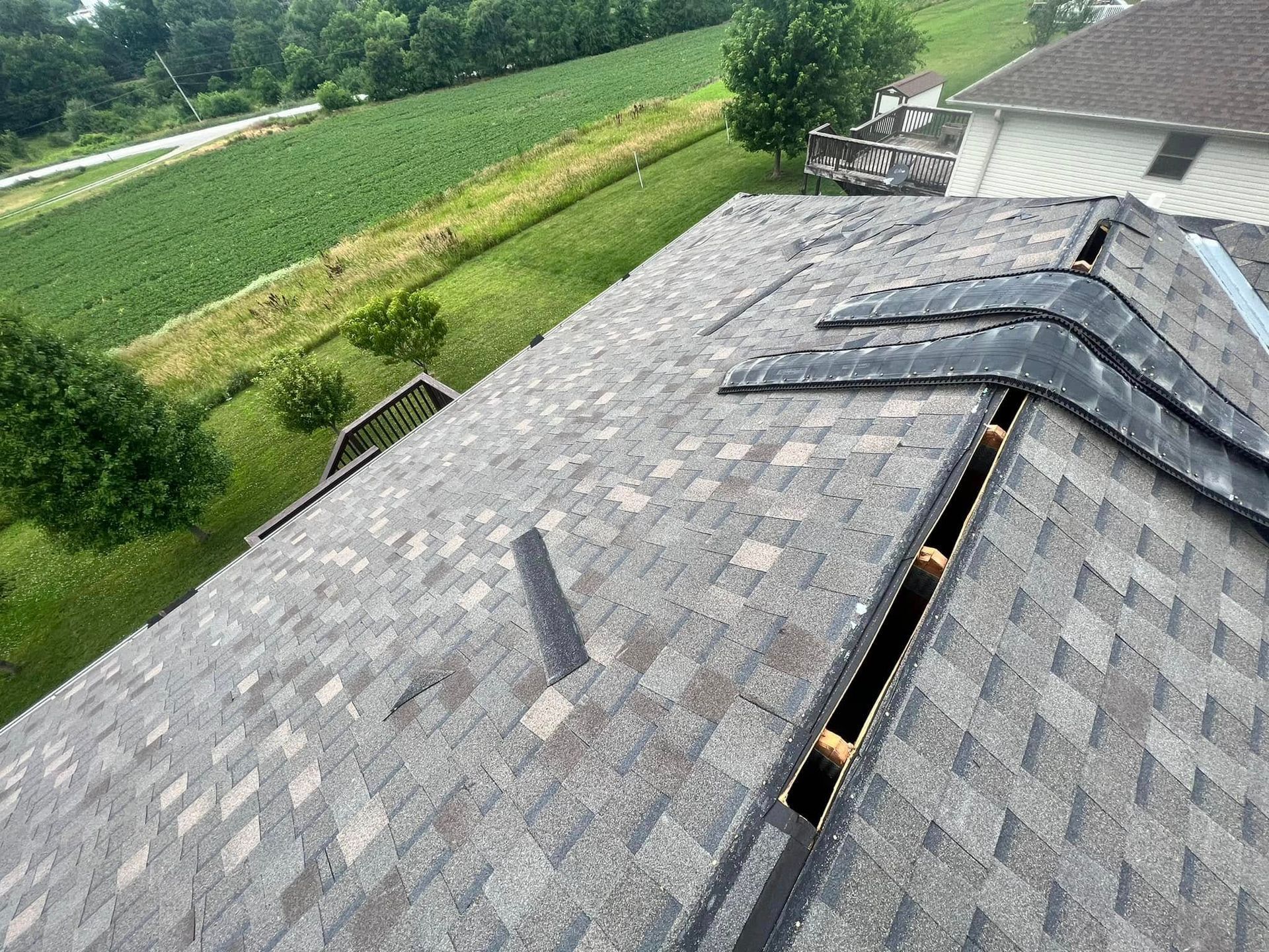 Damaged shingled roof with a long open gap near the ridge, overlooking green fields.