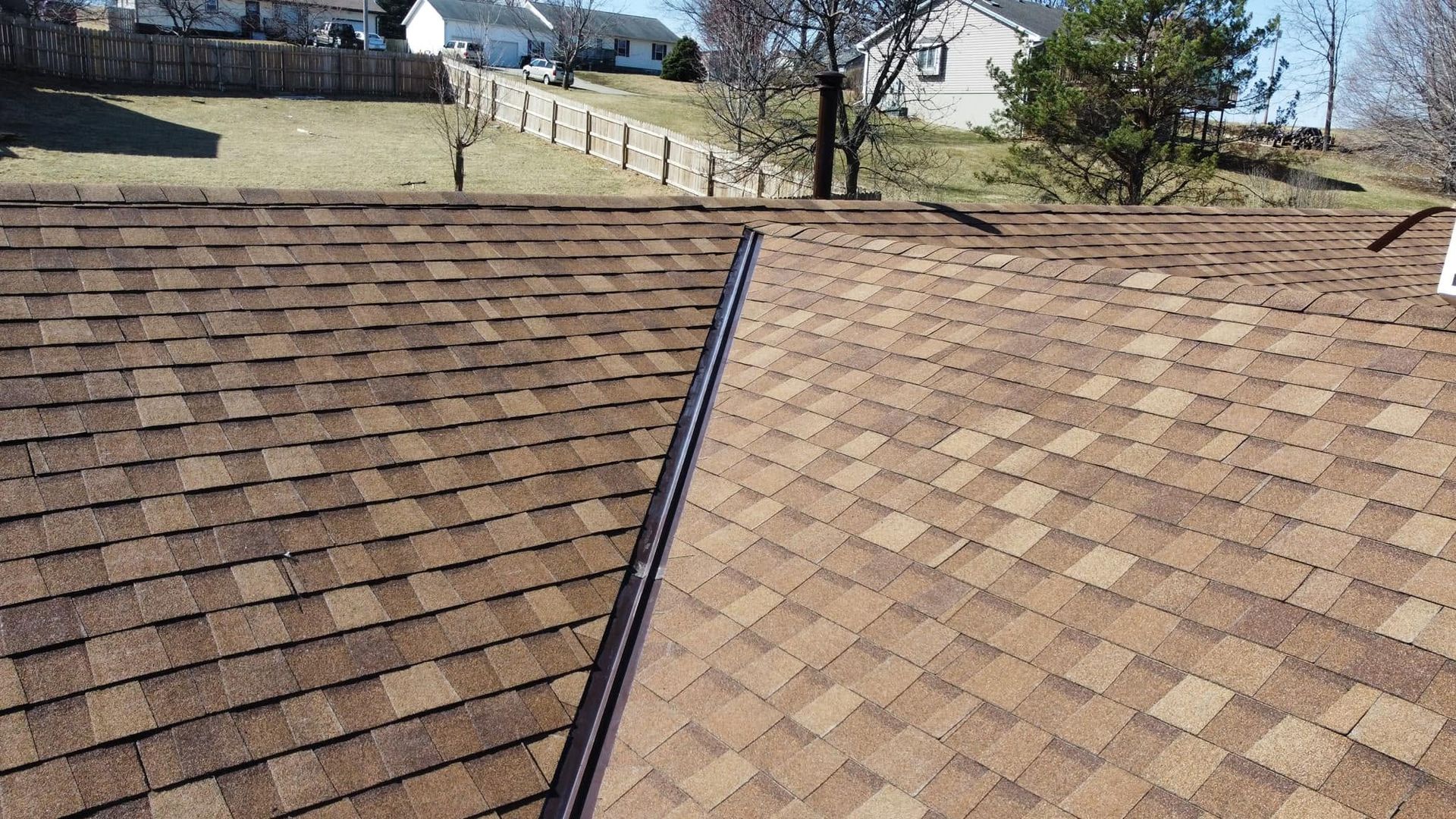 Brown shingle roof with a black ridge vent, viewed toward a backyard fence and trees