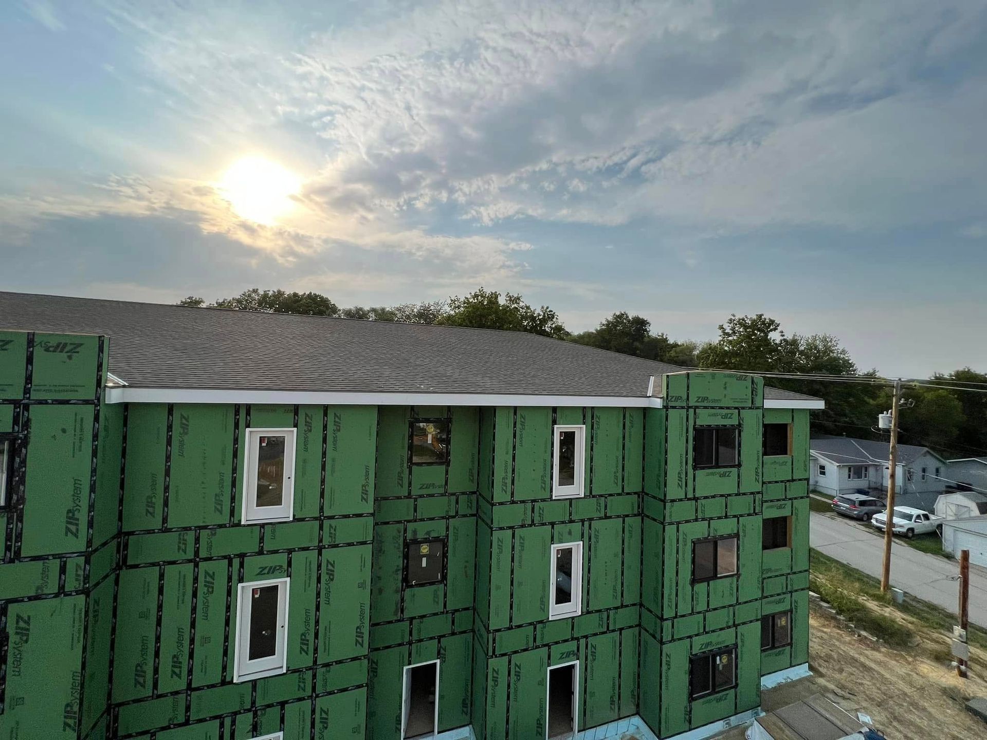 Green apartment building under construction at sunset, with exposed panels and a cloudy sky.