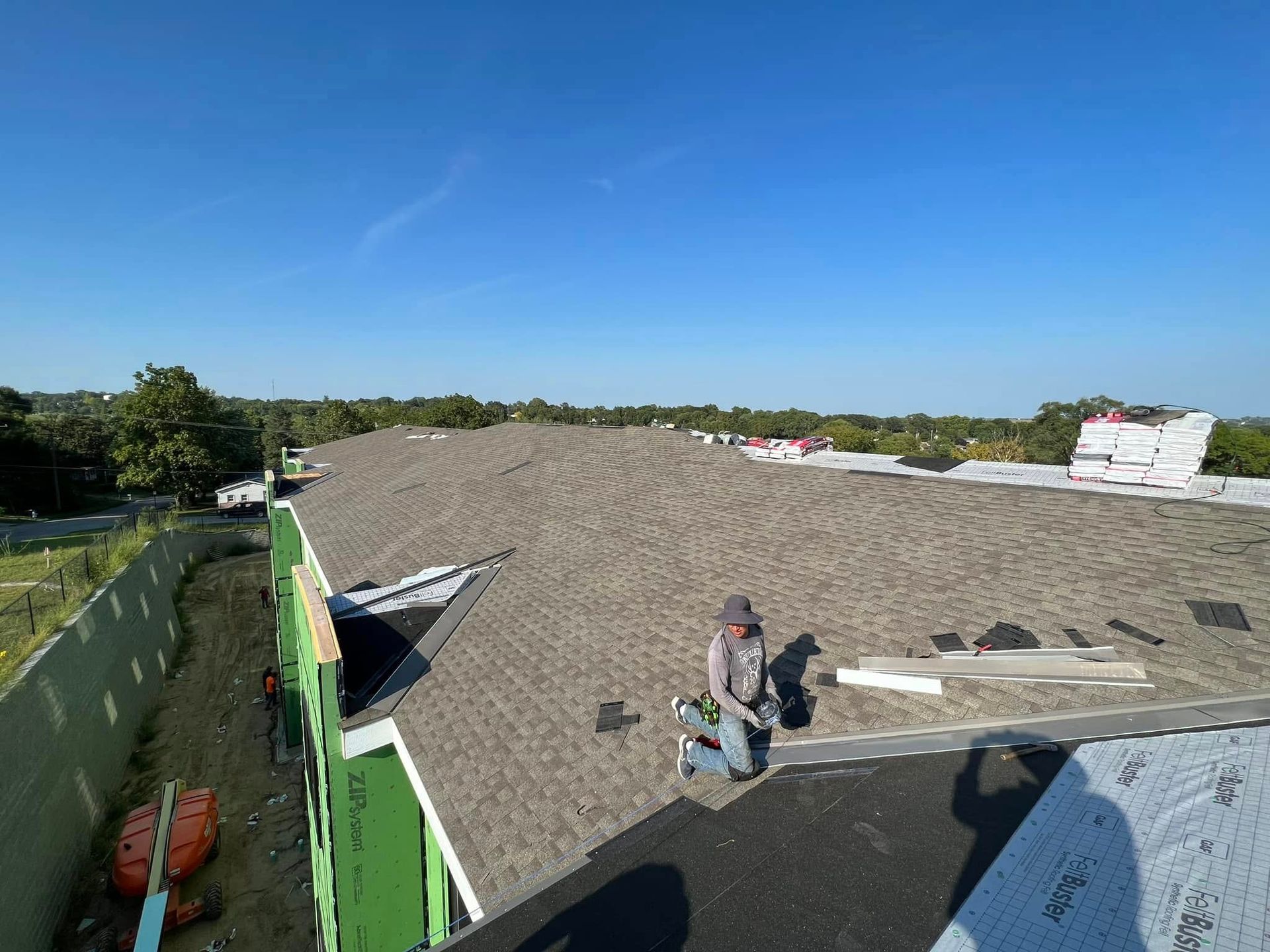 Workers on a roof under a clear blue sky, moving materials near a green wall.