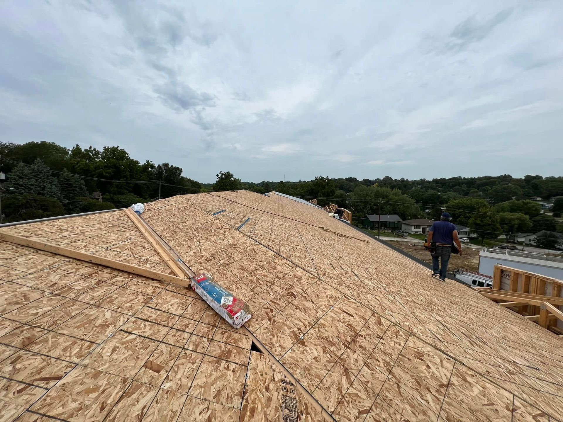Workers installing plywood roofing on a house under construction, with a cloudy sky and trees nearby
