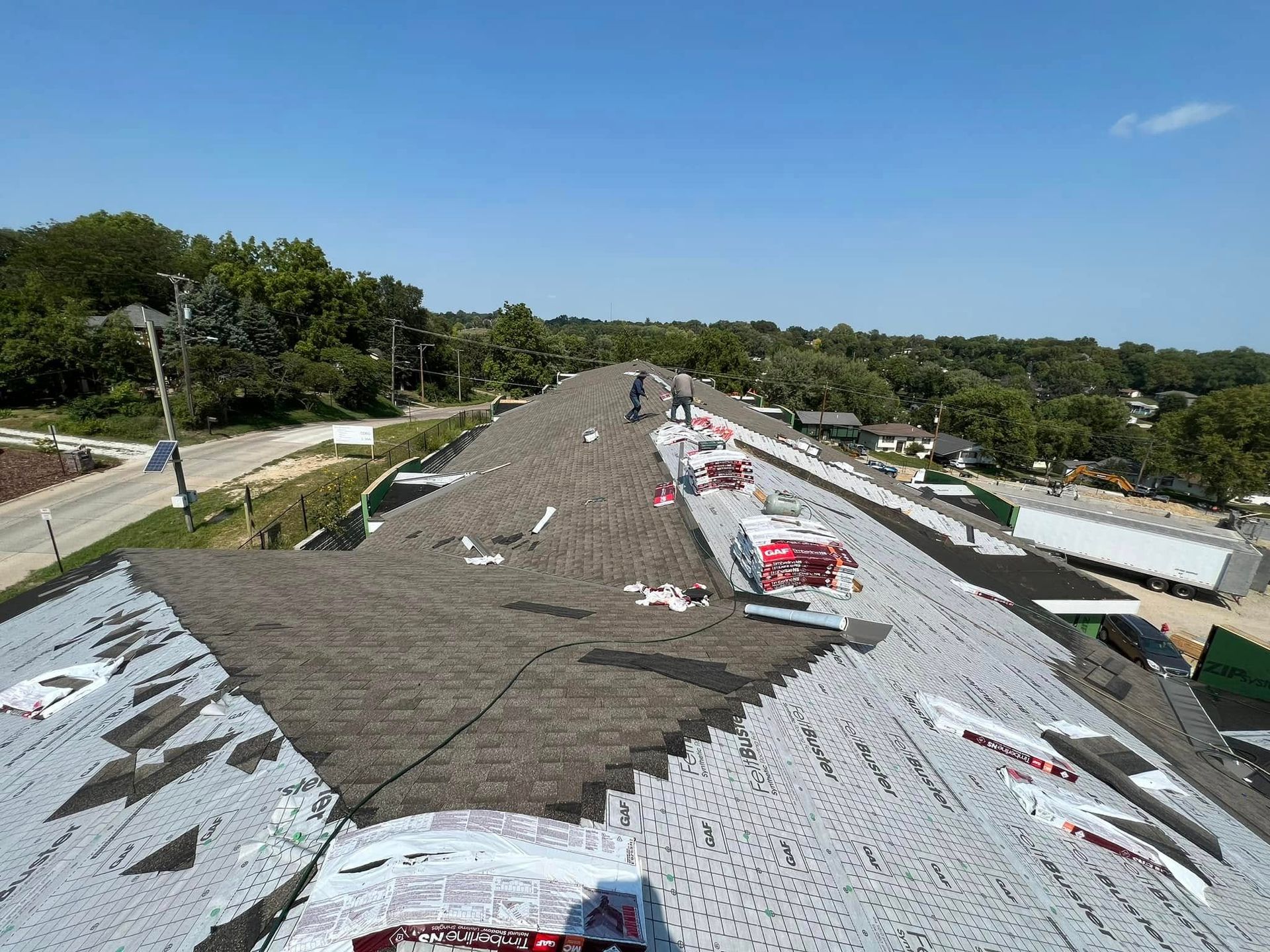 Roofer working on a shingled roof under a clear blue sky, with trees and buildings in the background