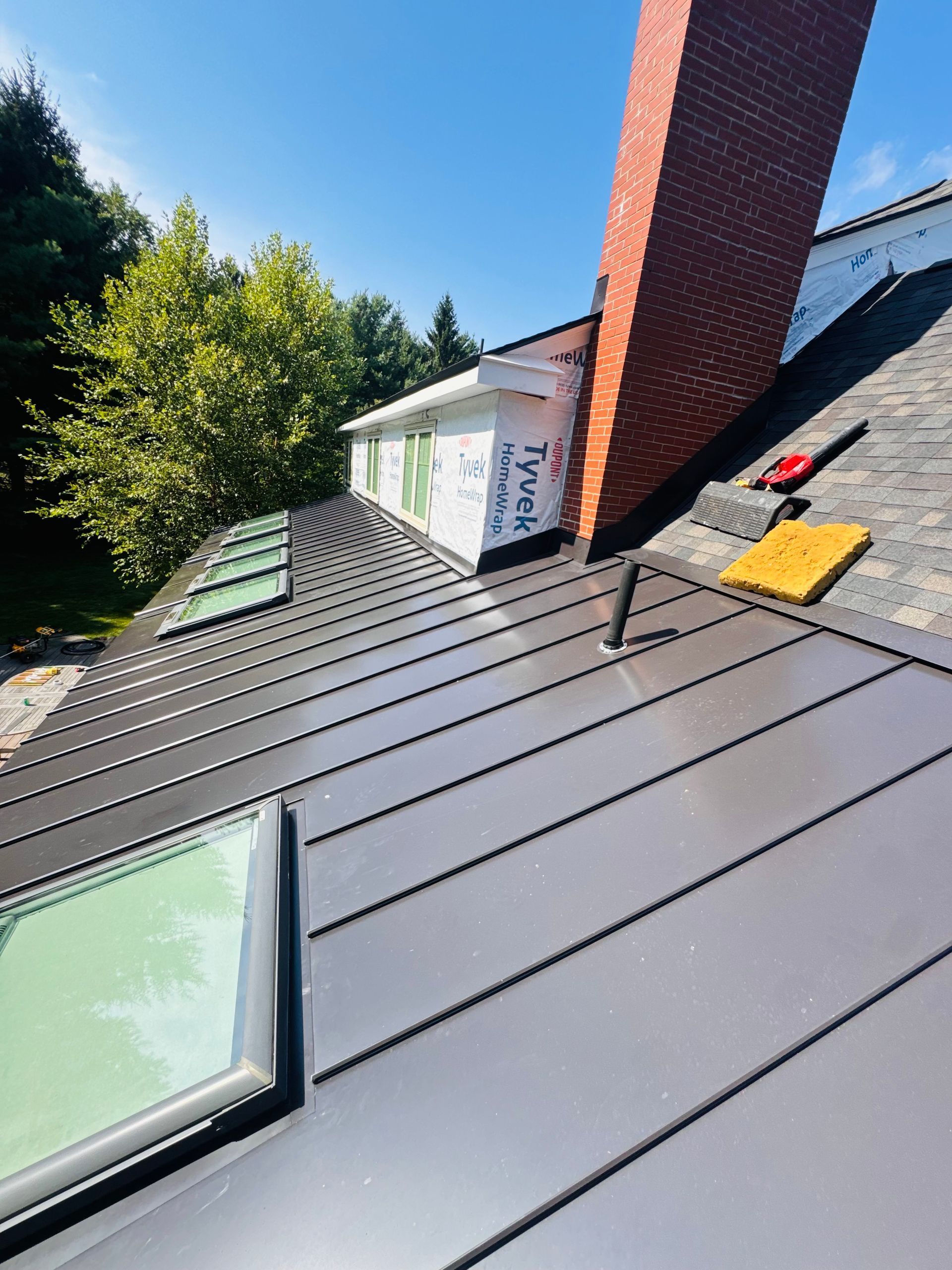 Sloped gray metal roof with skylight, red chimney, and yellow tool near the ridge under a blue sky