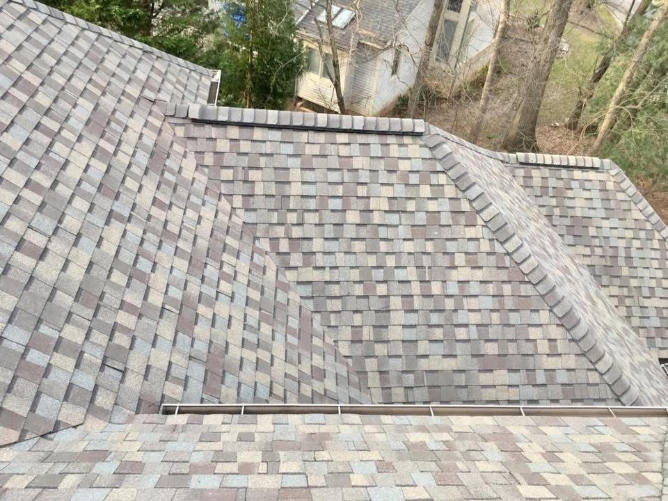 Gray shingled rooftop with valleys and vents, viewed from above, with trees in the background
