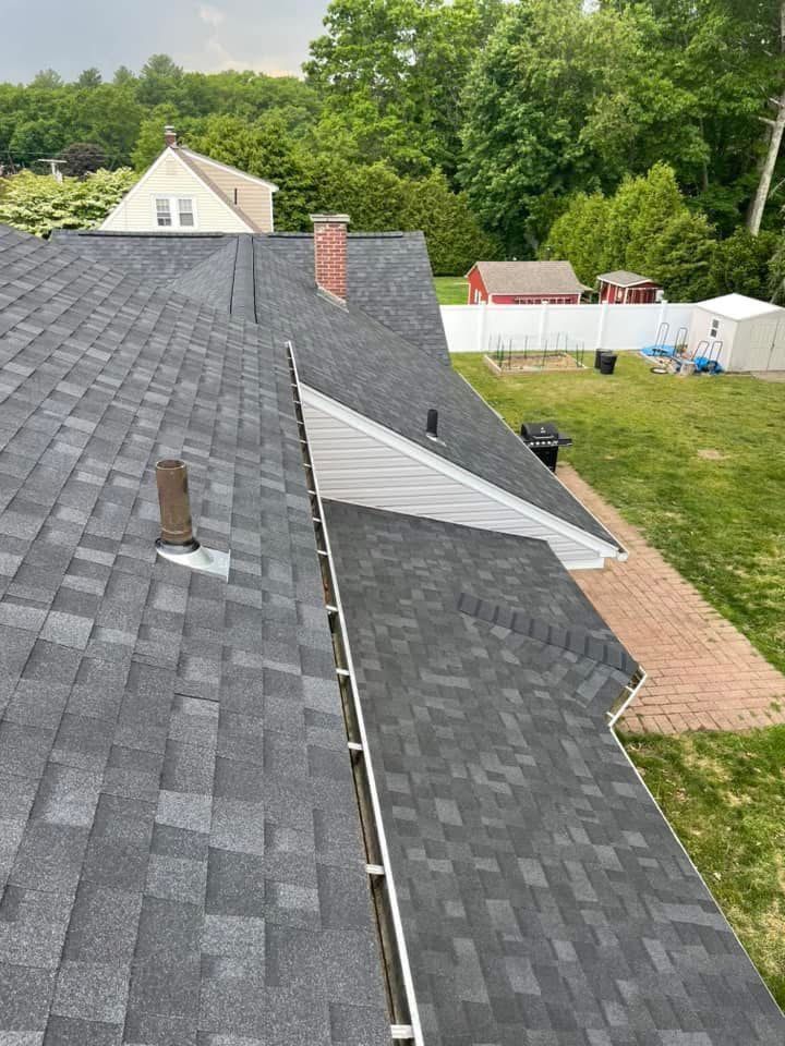 View from a shingled roof over a backyard with trees, a brick chimney, and white fencing.
