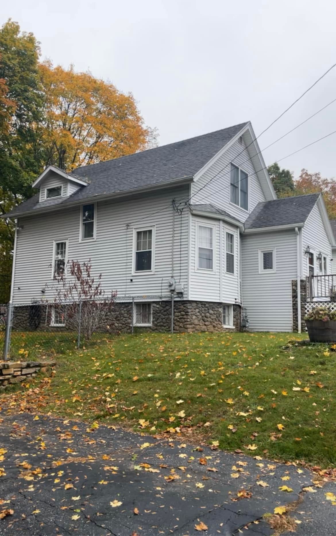 Gray two-story house with white trim and autumn leaves on the lawn beside a wet street