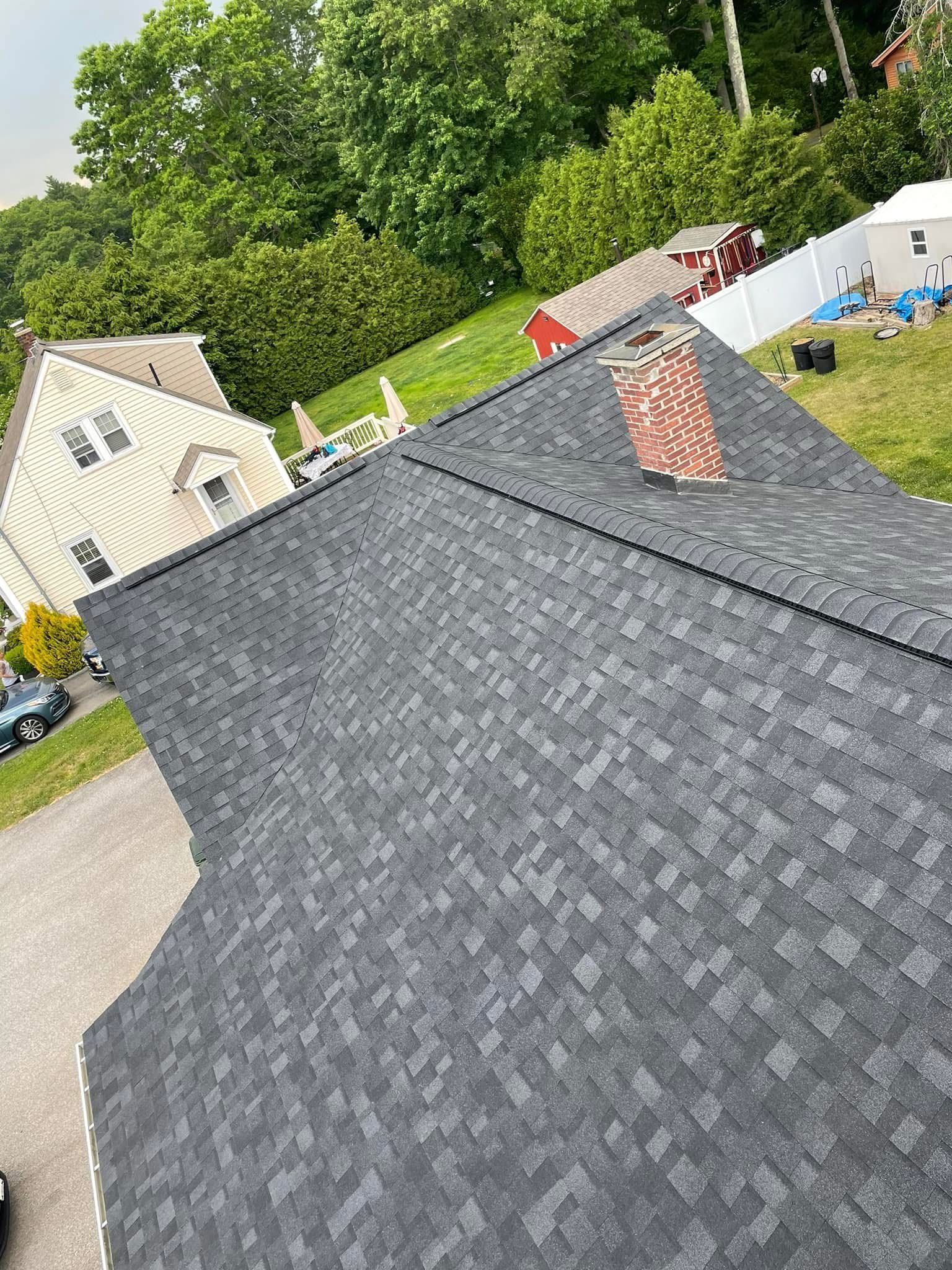 Gray shingled roof with a brick chimney in a suburban neighborhood, trees and houses in the background