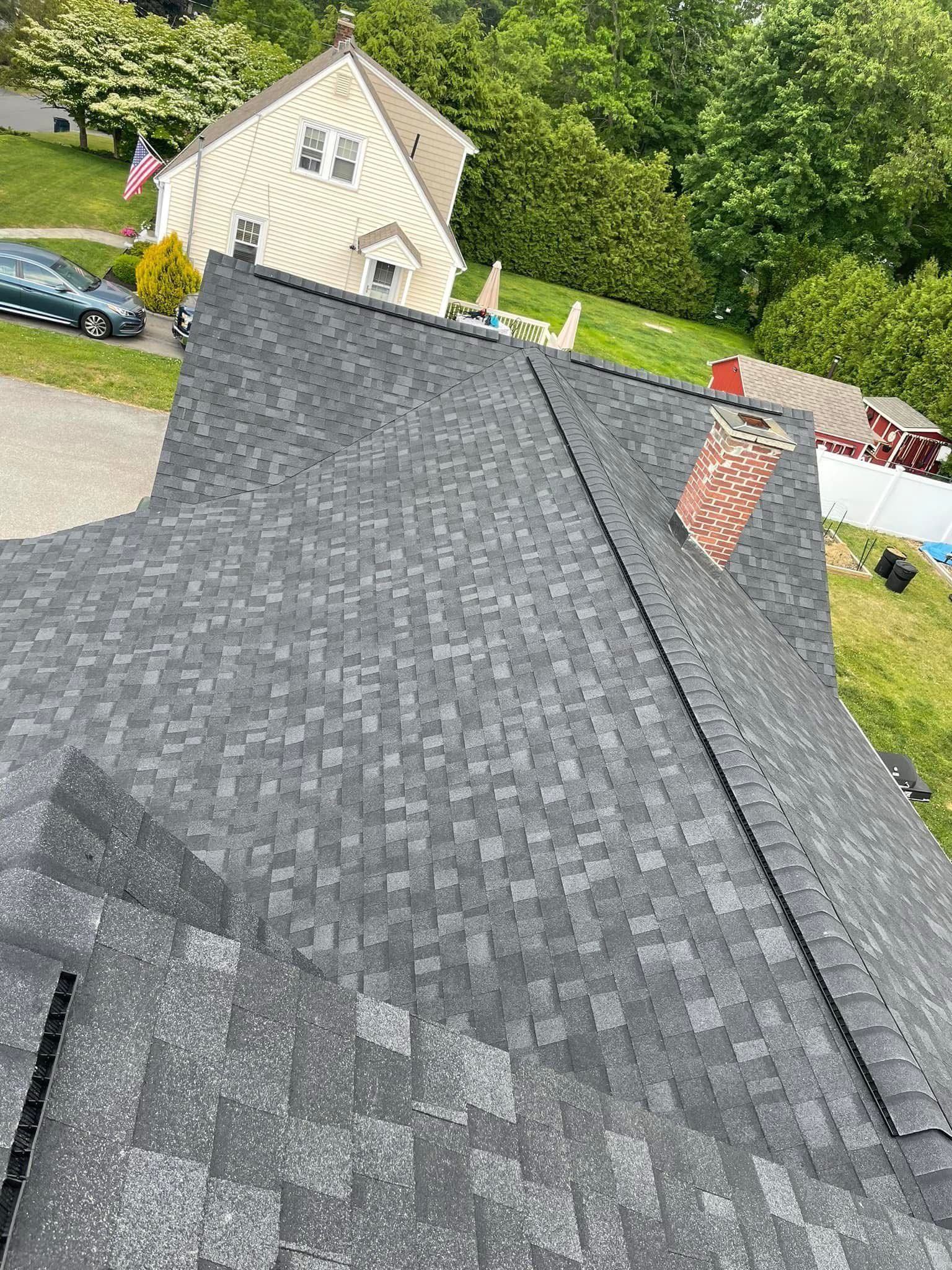 Roof with dark gray shingles and a small chimney, viewed from above with a house and trees in the background