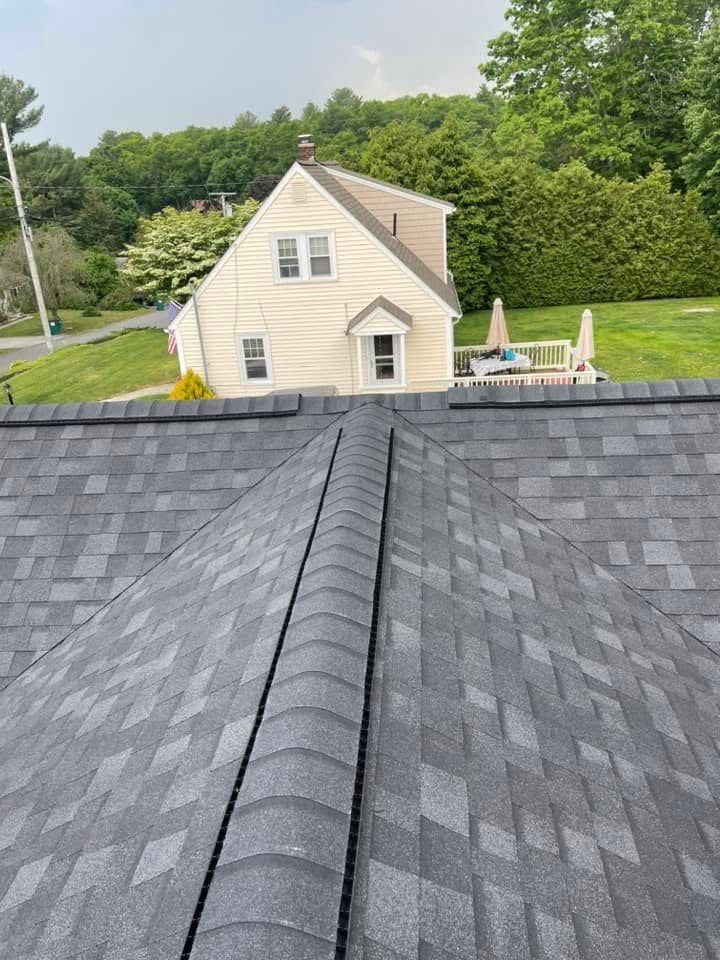 Gray shingle roof in foreground with a white house and green trees in the background