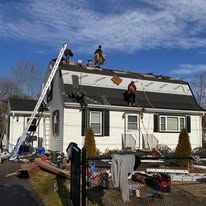 Workers on a roof and ladder repairing a white house with roof damage and debris in the yard
