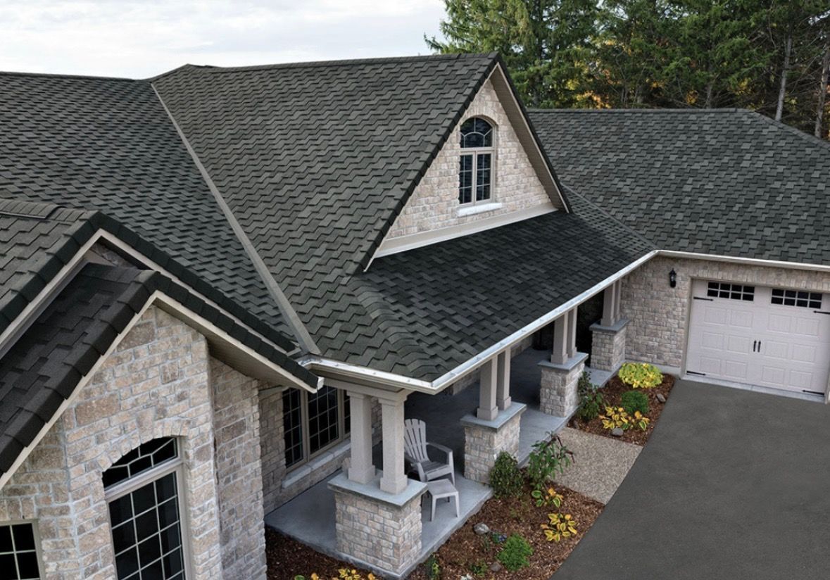 Aerial view of a stone house with dark shingle roof, front porch, and attached garage