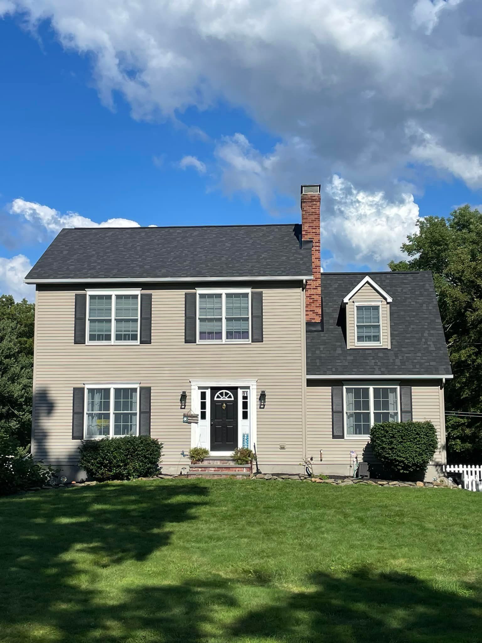 Two-story beige house with dark roof, brick chimney, and front lawn under a blue sky.
