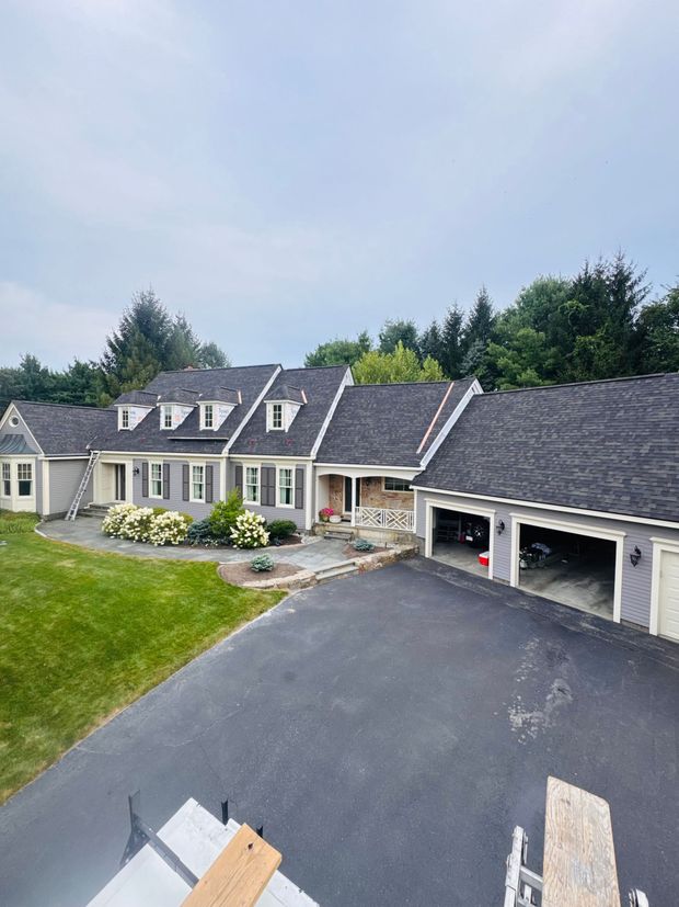 Aerial view of a suburban house with dark roof, attached garage, paved driveway, and green lawn.