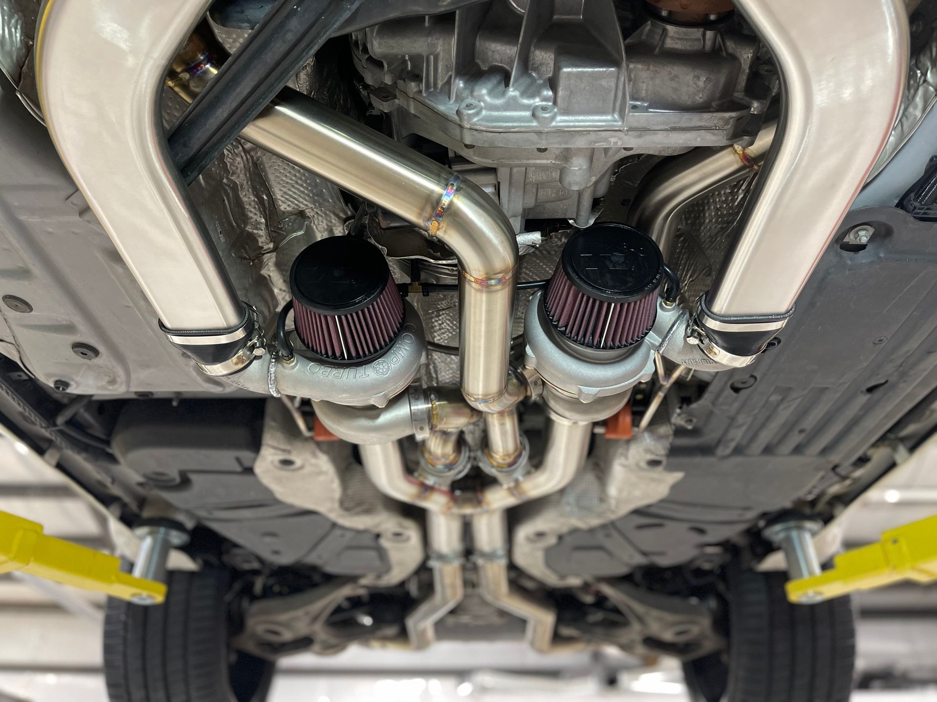 Underneath a car with a custom exhaust system. Two turbos with red air filters, chrome pipes, and yellow jack stands.