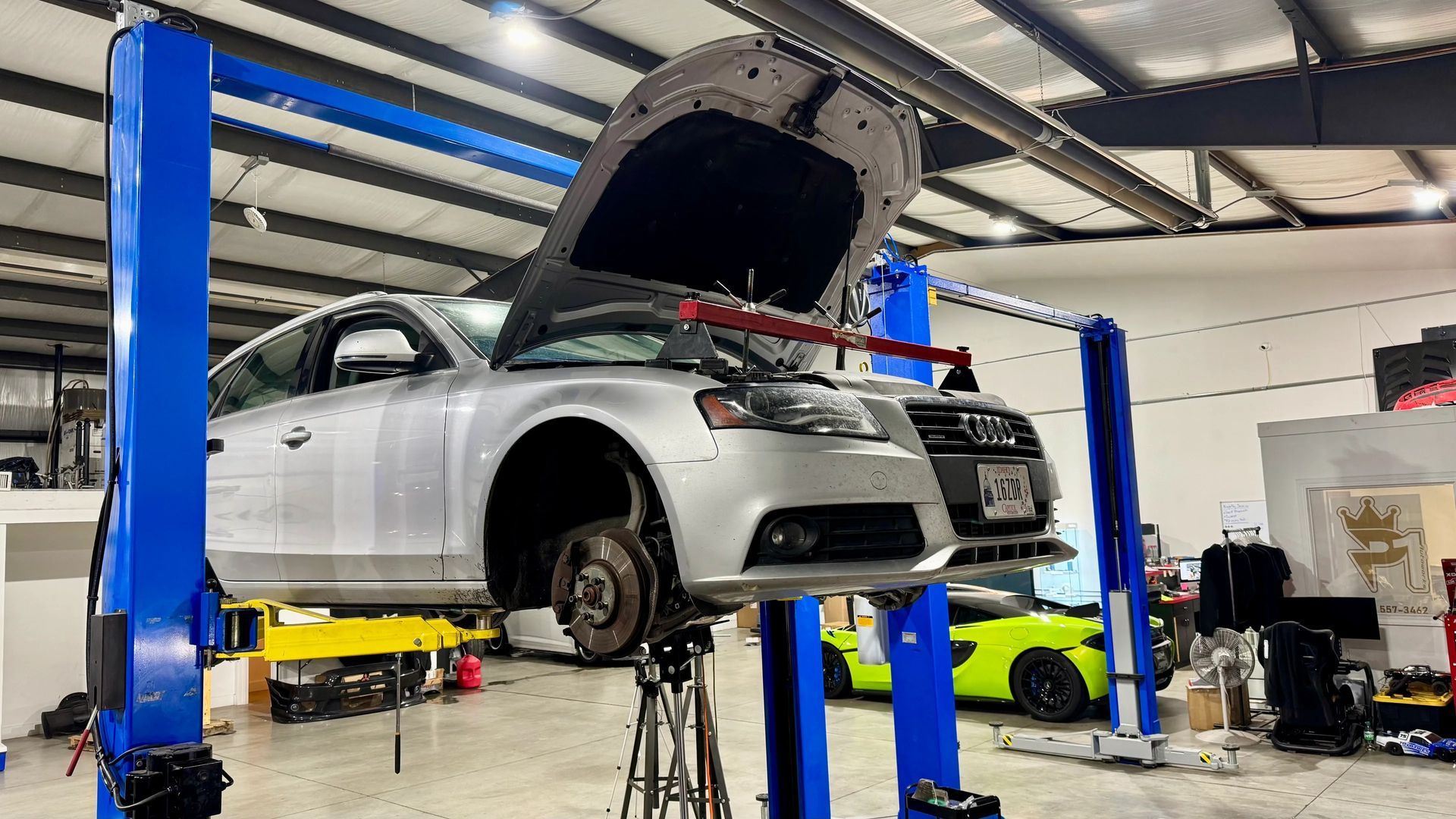 Silver car raised on a lift in a garage with hood open, wheel off, and yellow car visible.