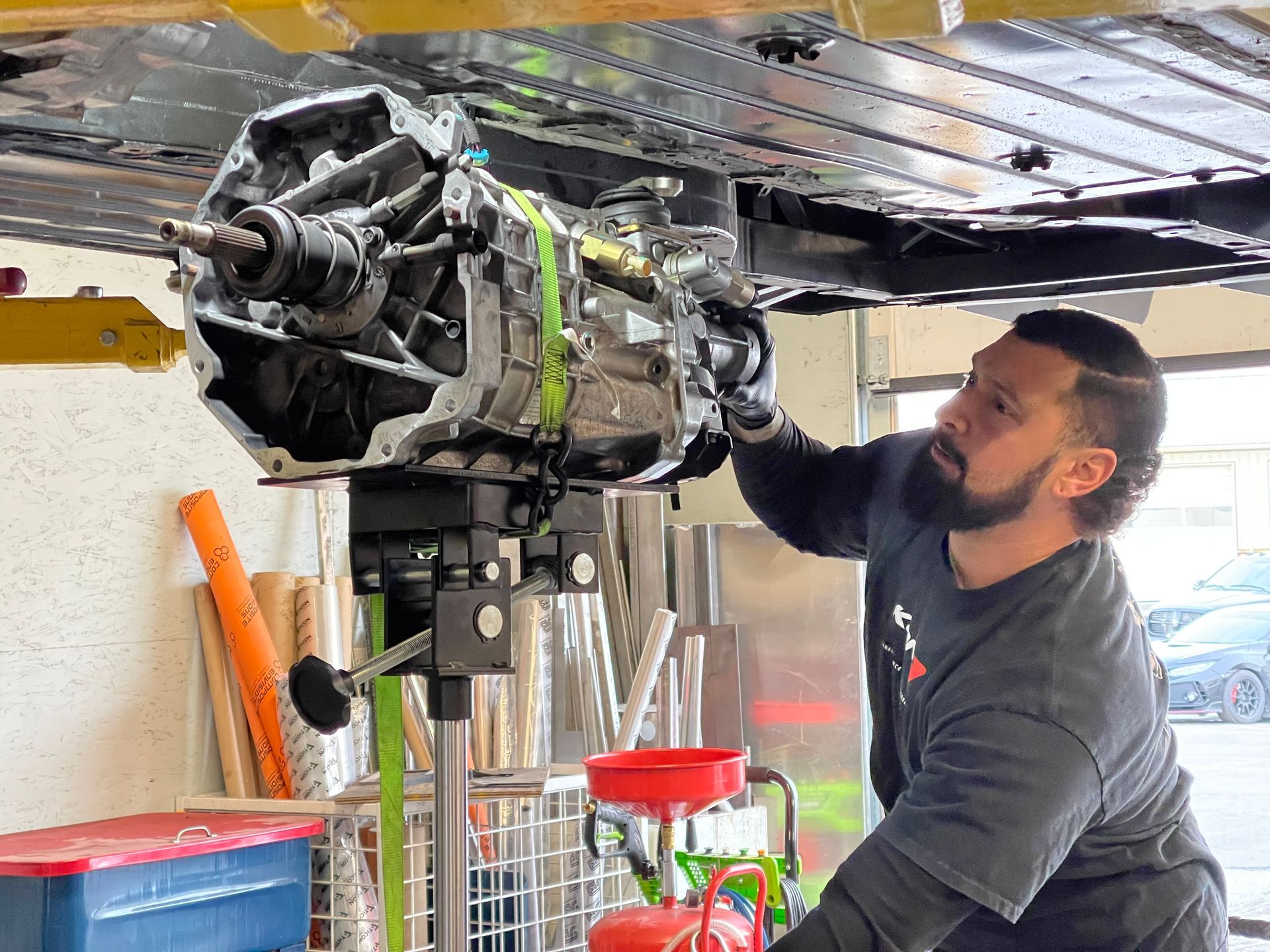 Mechanic using a transmission jack to remove a car's transmission. Garage setting, sunlight, red fluid collection.