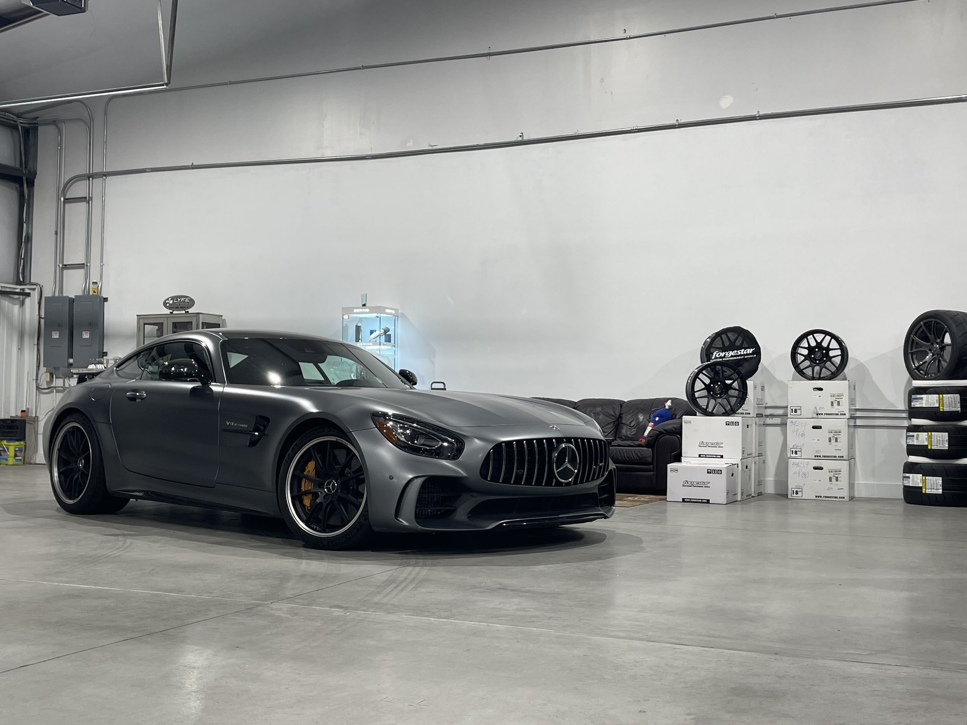 Gray sports car in a garage with black rims, boxes, and tires against a white wall.