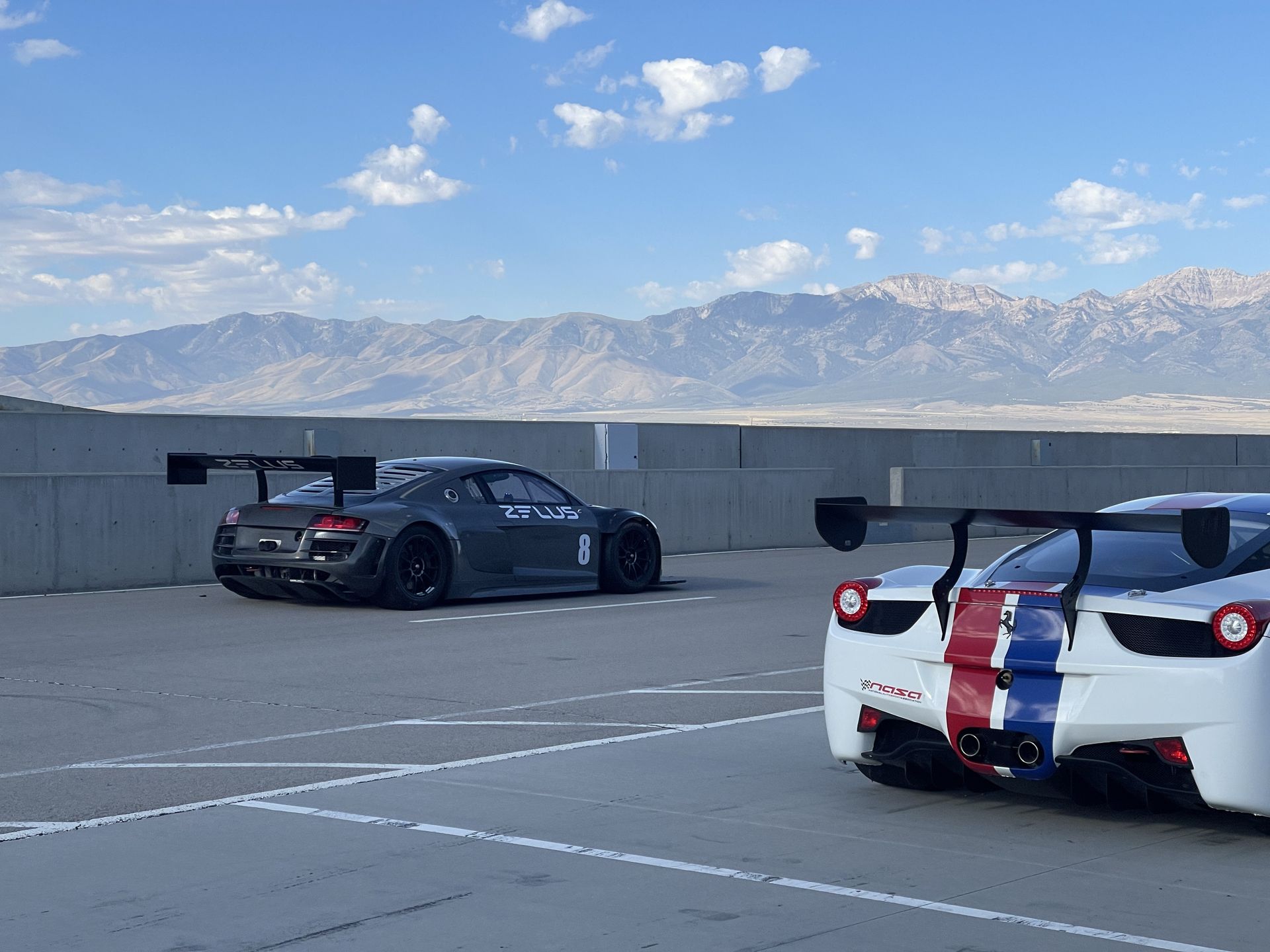 Two race cars on a parking lot, one gray Audi R8, the other white Ferrari with blue/red stripes. Mountains in the background.