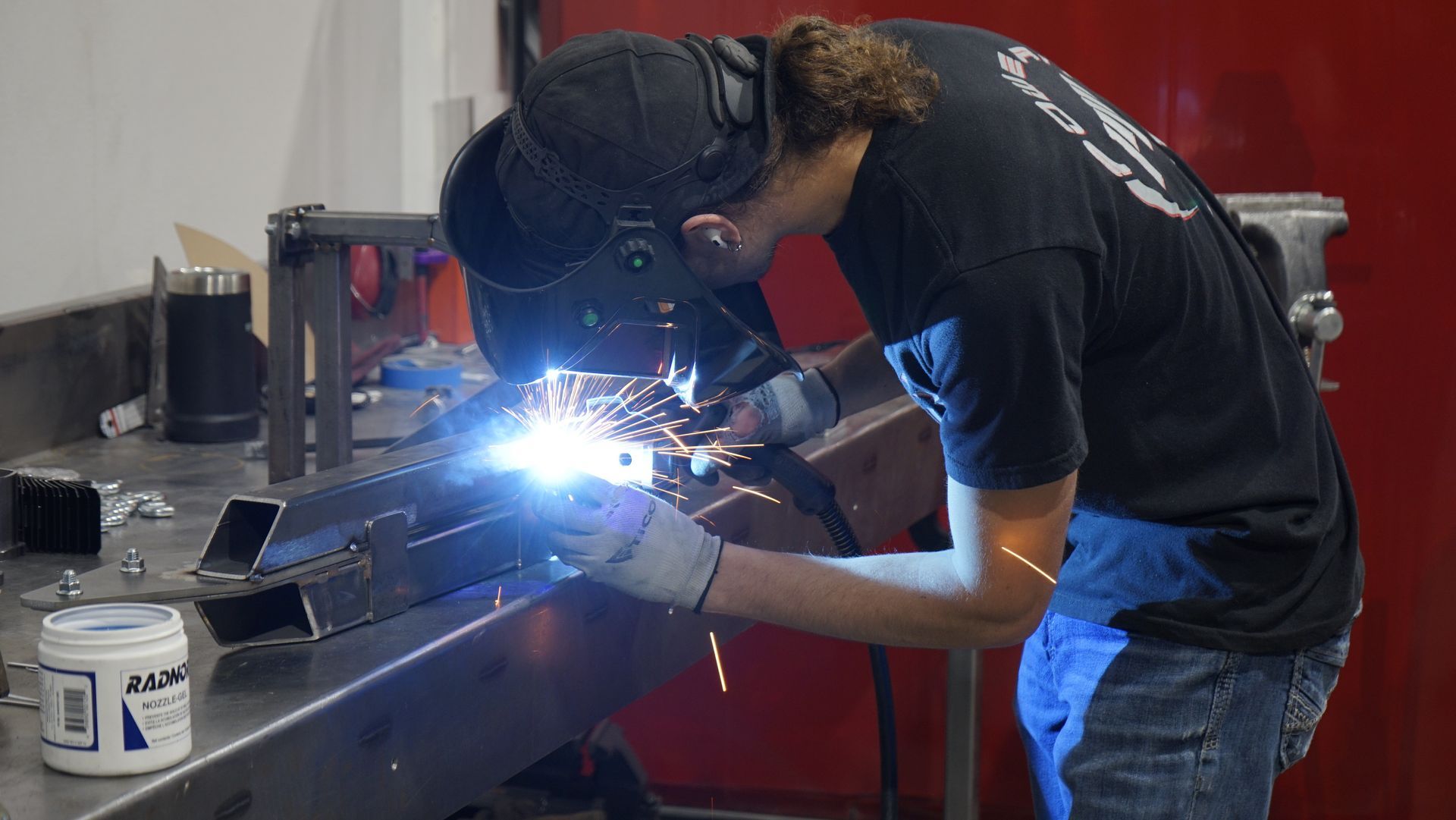 Person welding metal in a workshop, wearing a welding helmet and gloves.