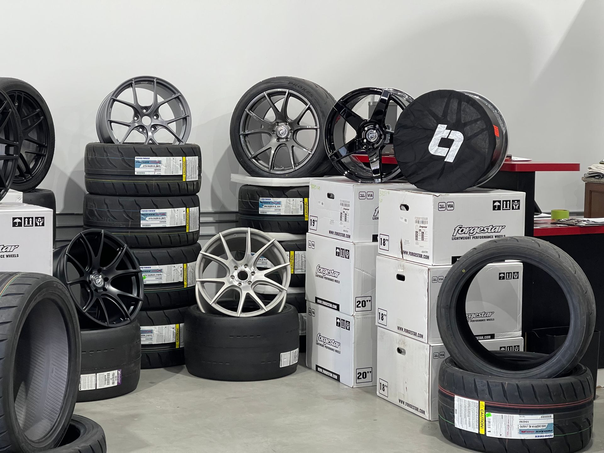 Tires and alloy wheels displayed in a warehouse setting. Various sizes and colors are visible, stacked for sale.