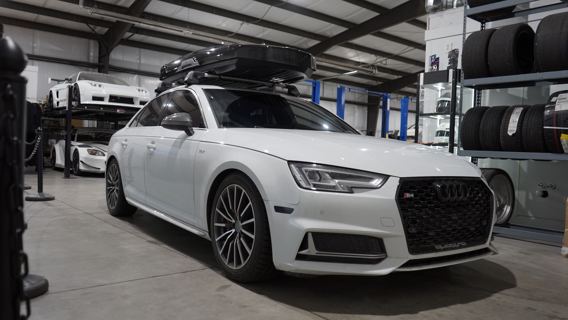 White Audi sedan with roof cargo carrier parked inside a garage, other cars and tires in background.