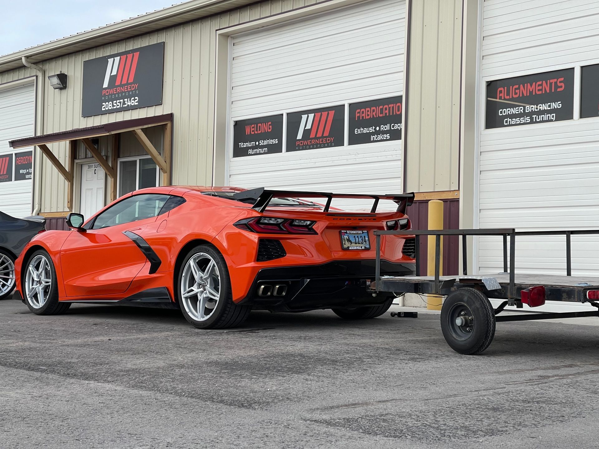 Orange sports car towing an empty trailer in front of a garage.