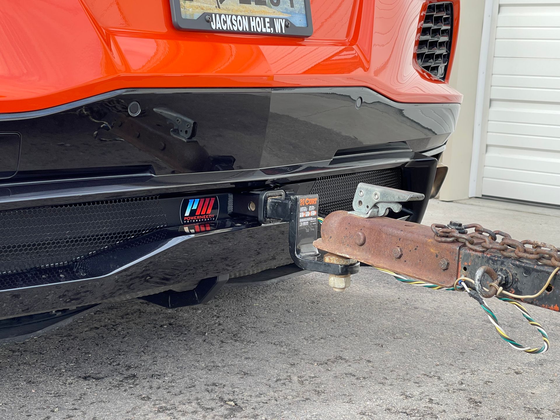 Rear of an orange sports car with a black bumper and trailer hitch connected to a rusty trailer.