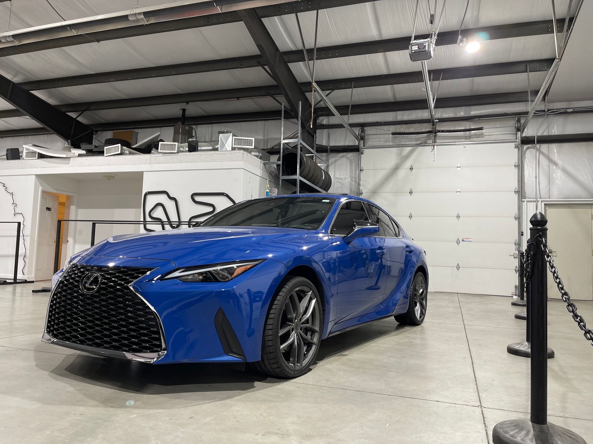 Blue Lexus coupe parked inside a garage with black ceiling beams and a white wall.