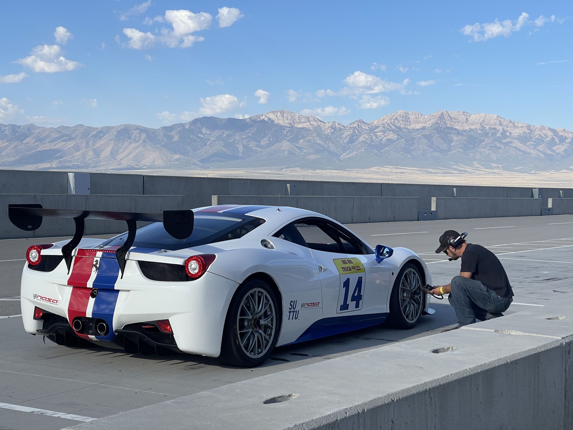 White race car with blue and red stripes on a track; a person kneels near a tire. Mountains in the background.