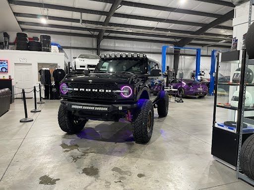 Black lifted Bronco with purple accents in a garage, next to other vehicles and displays.