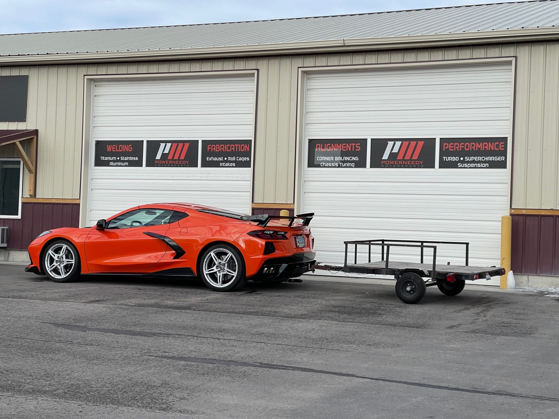 Orange sports car towing a small trailer in front of a garage with 