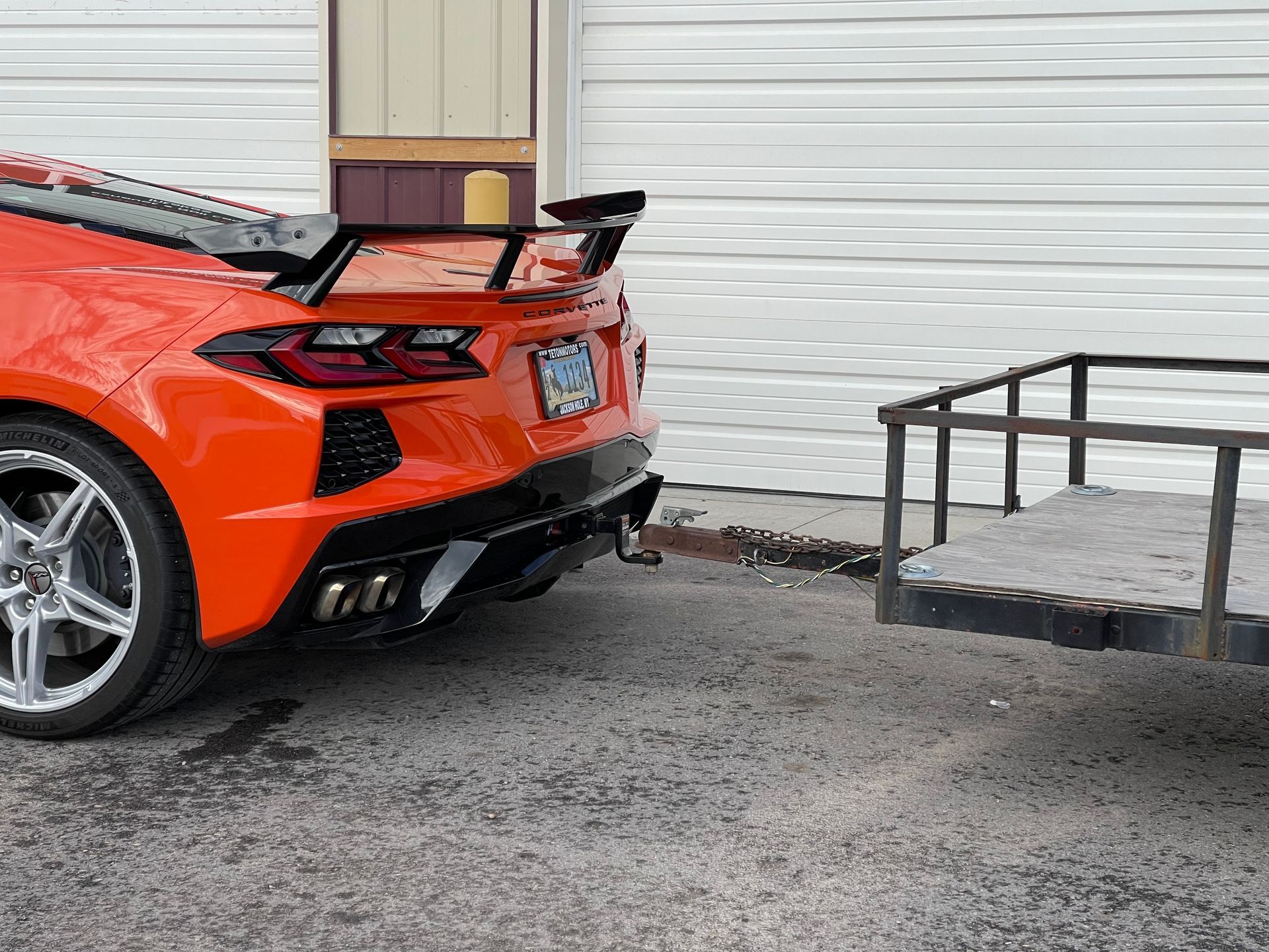 Orange sports car towing an empty trailer outside a building.