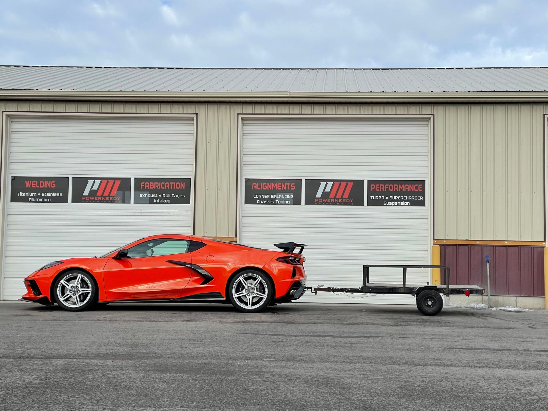 Orange sports car pulling a trailer in front of a building with garage doors.