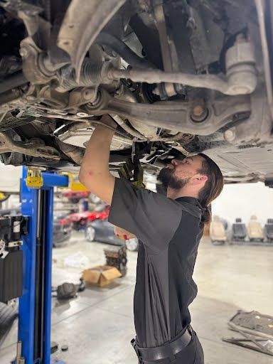 Mechanic working on the undercarriage of a car in a shop. He is reaching up toward the vehicle.