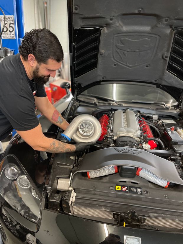 Man working on a car engine, installing a turbocharger. Hood open, black car, garage setting.