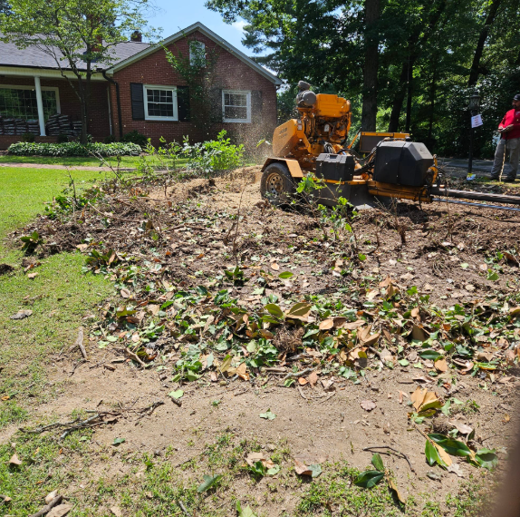 A stump grinder removes a tree stump in a yard. Debris and a brick house are visible.