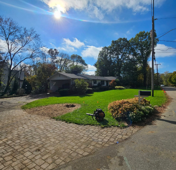 Ranch-style home with green lawn, blue sky, and a person using a leaf blower on the grass.