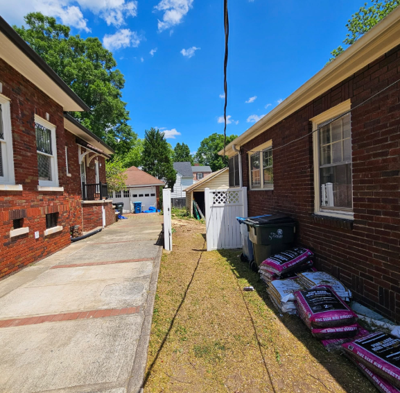 Brick buildings flank a narrow alley, with a driveway on the left and bags of mulch on the right. Blue sky overhead.