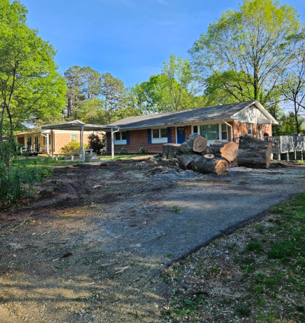 A ranch-style house with cut logs in the front yard. Dirt and gravel driveway in front.