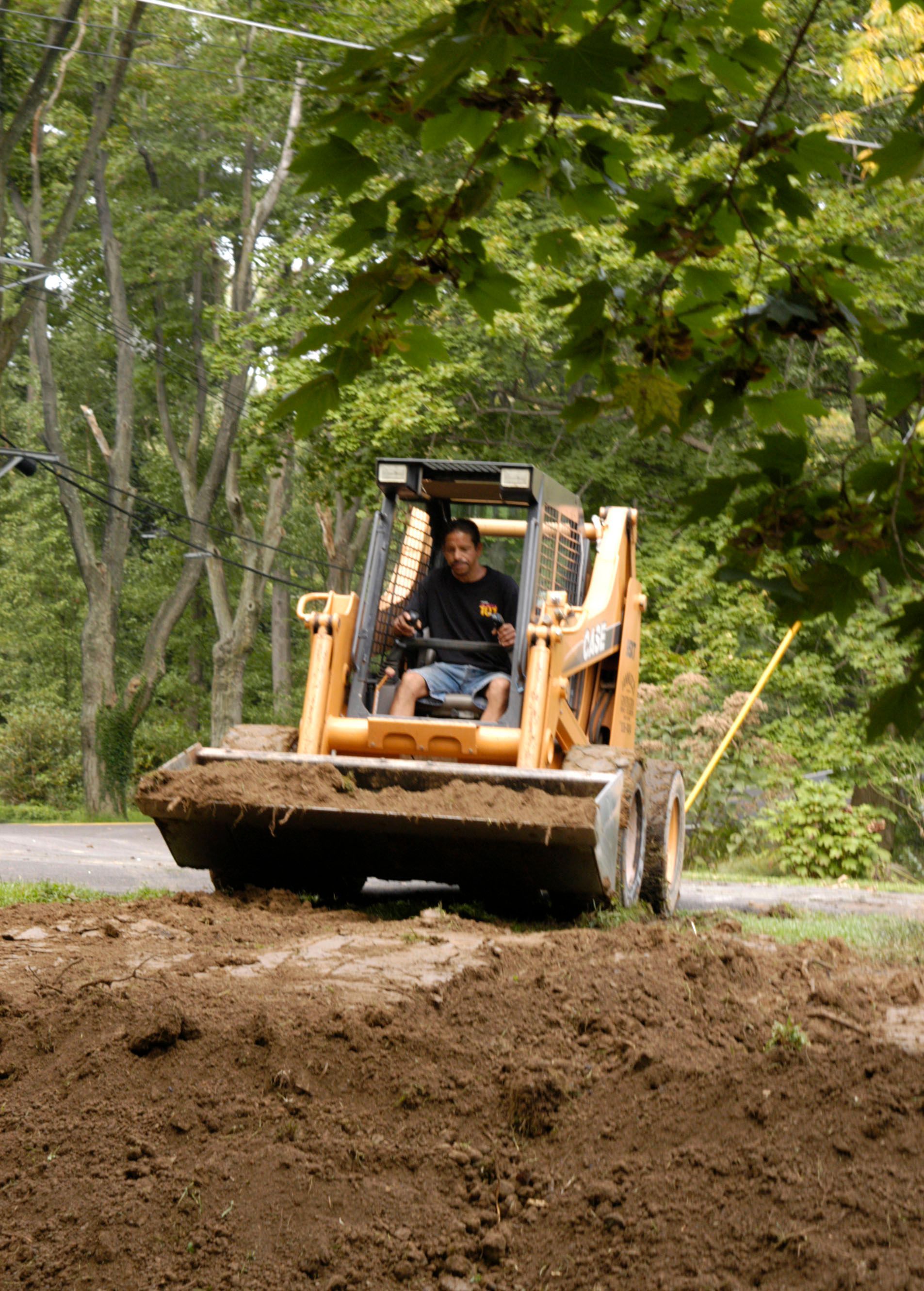Man operating a skid steer, pushing dirt on a roadside. Trees and foliage in background.