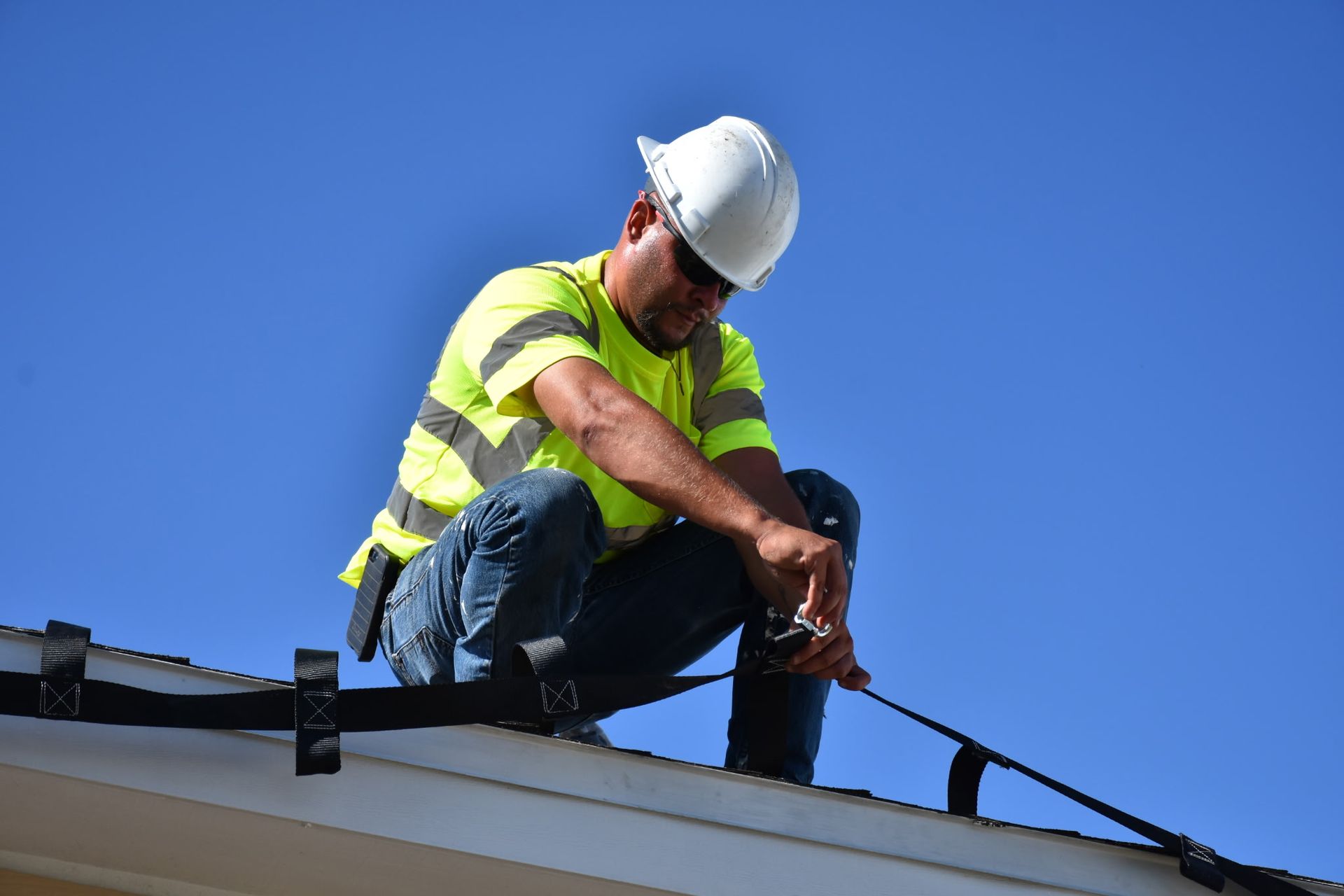 Construction worker on a roof wearing a safety vest and helmet, securing equipment against a blue sky.