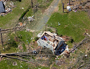 Aerial view of a house damaged by a storm, with roof partially missing, surrounded by fallen trees and debris.