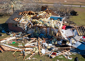 Destroyed house with debris scattered across the yard after a storm. Roof and walls are collapsed.